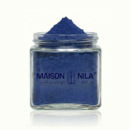 Un pot en verre transparent de 100 g de Poudre de Nila, rempli de poudre bleue vibrante et étiqueté MAISON NILA en anglais et en arabe. Le pot ouvert montre la poudre bleue distinctive amassée au-dessus du bord.